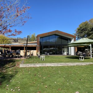 Places To Go Auckland 4 Modern building with large windows on grassy lawn, tables with umbrellas, trees, mountains visible.